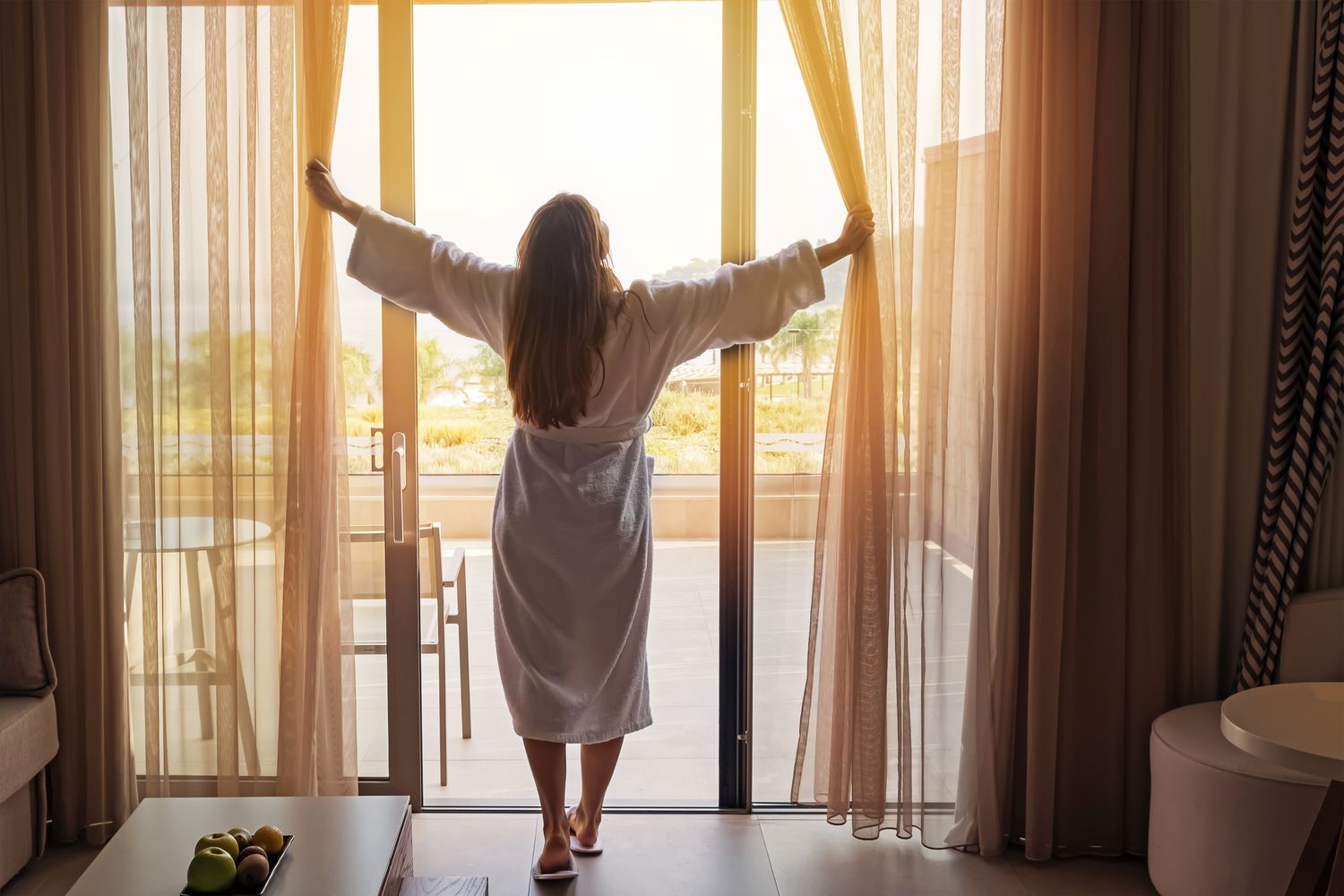 Woman in robe opening curtains to sunlight.