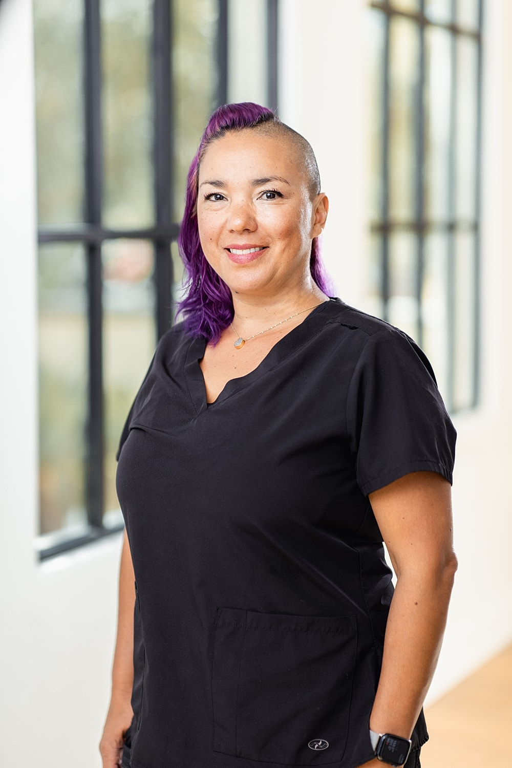 Smiling woman with purple hair in black scrubs.
